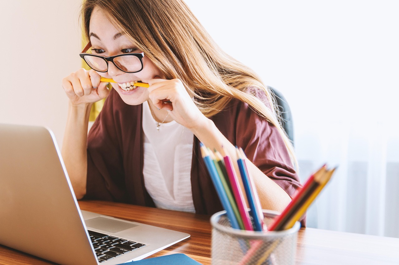 A woman in glasses bites a yellow pencil while looking at a laptop screen with a stressed expression.