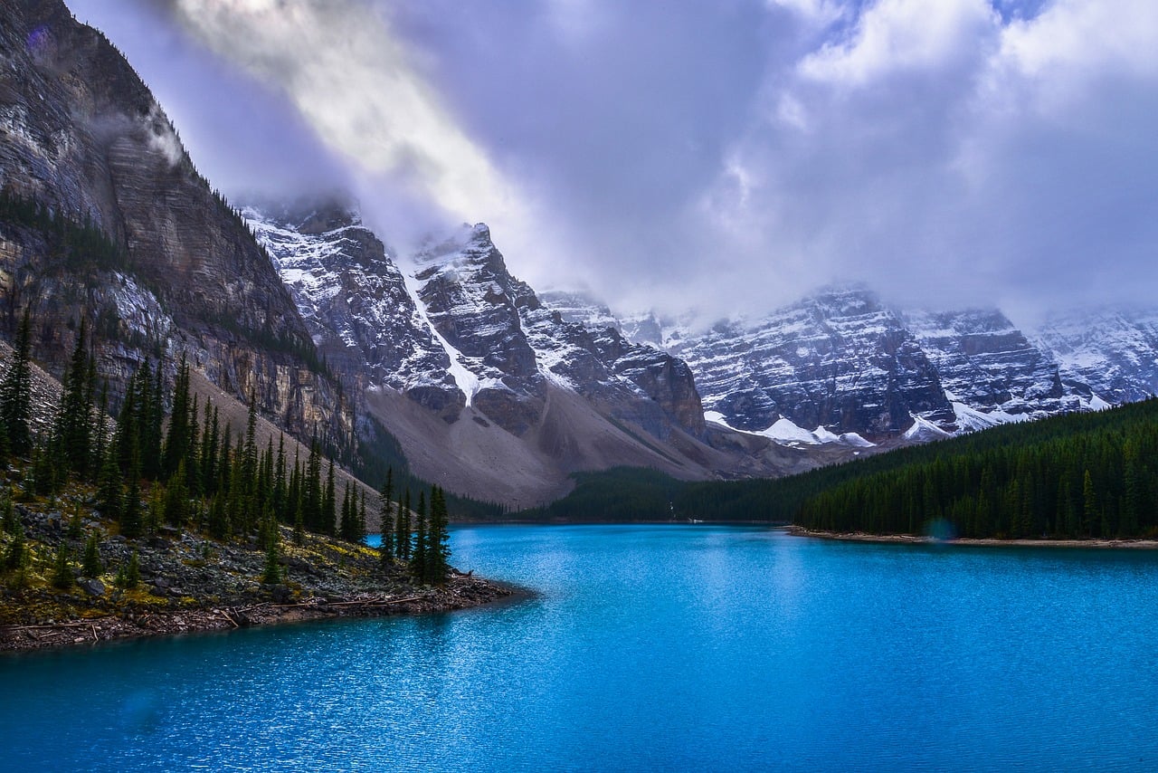 Vibrant blue lake surrounded by snow-capped mountains and dense green pine forests under a cloudy sky.