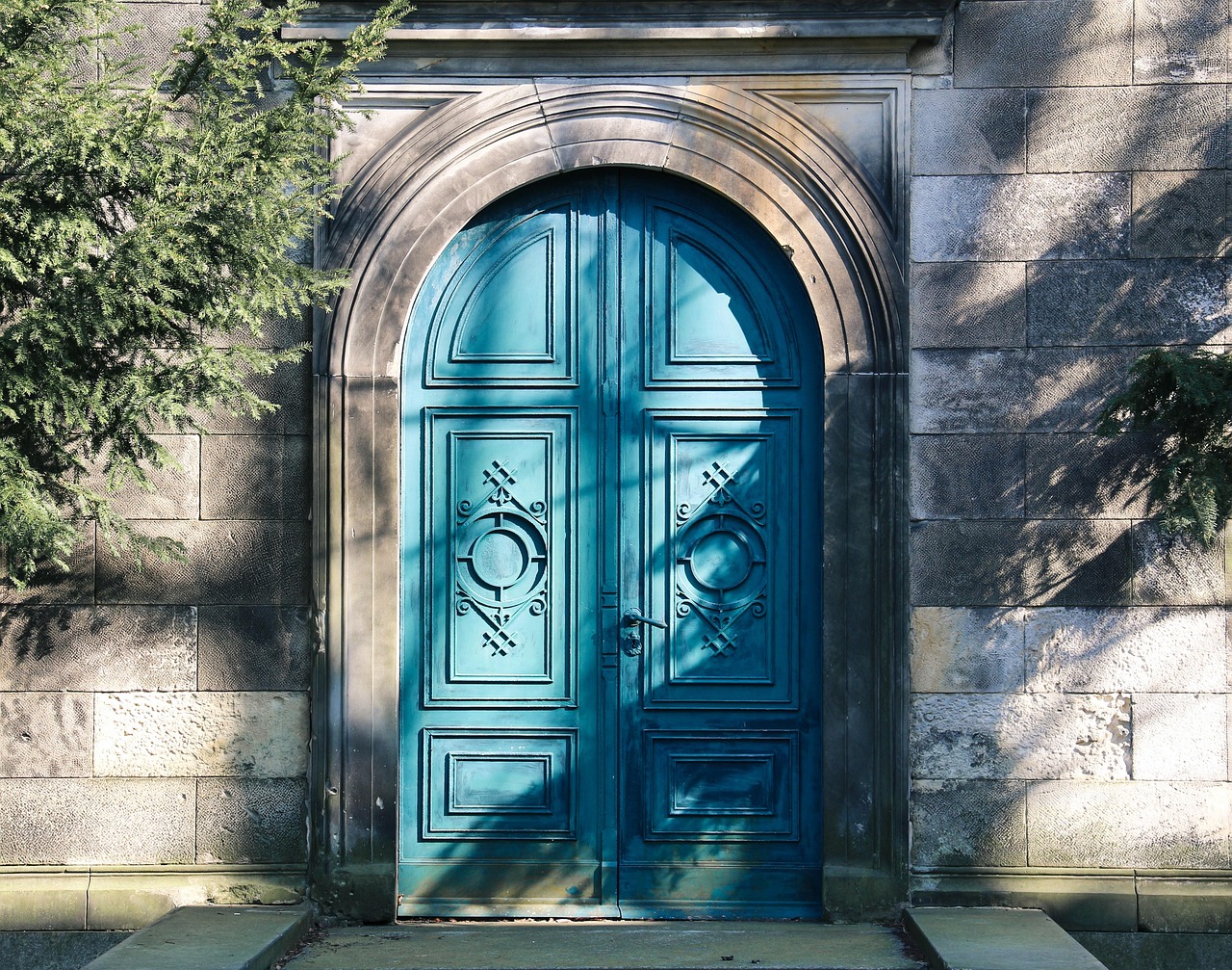 Ornate dark teal double door with decorative panels set into a stone wall, with dappled sunlight and tree foliage.