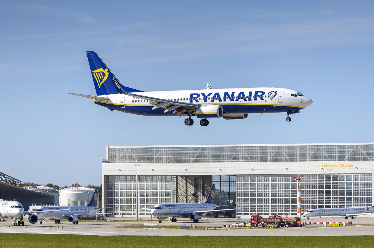 A Ryanair airplane flying low over an airport with hangars and other parked aircraft on the tarmac.
