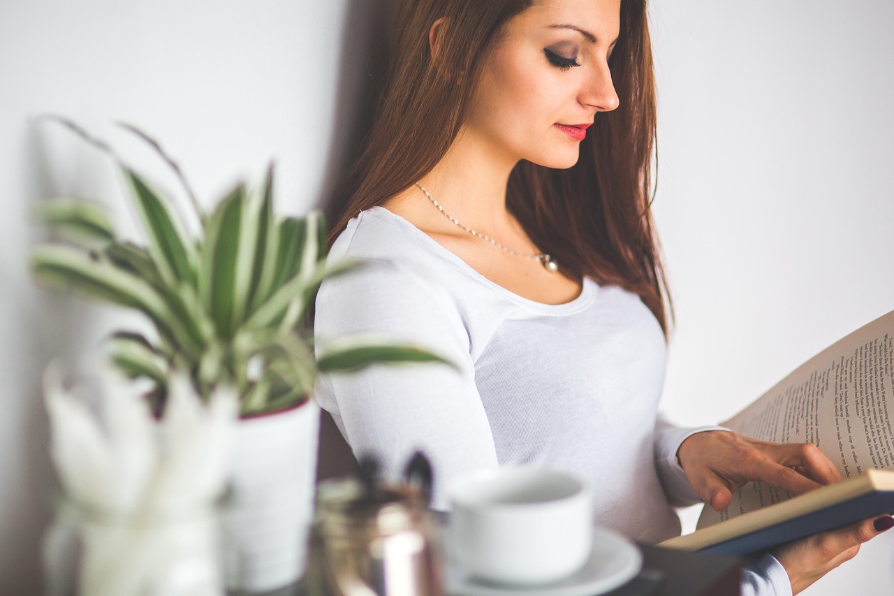 A woman with long brown hair wearing a white top and necklace, looking down and reading an open book.