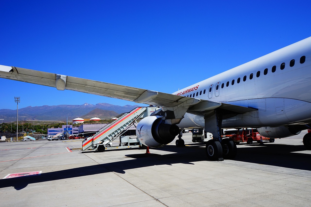 White passenger airplane with mobile boarding stairs on an airport tarmac, mountains in background.