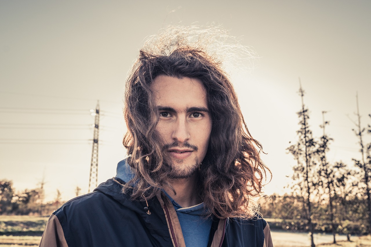 Close-up portrait of a young man with long wavy brown hair and a beard, wearing a dark jacket.