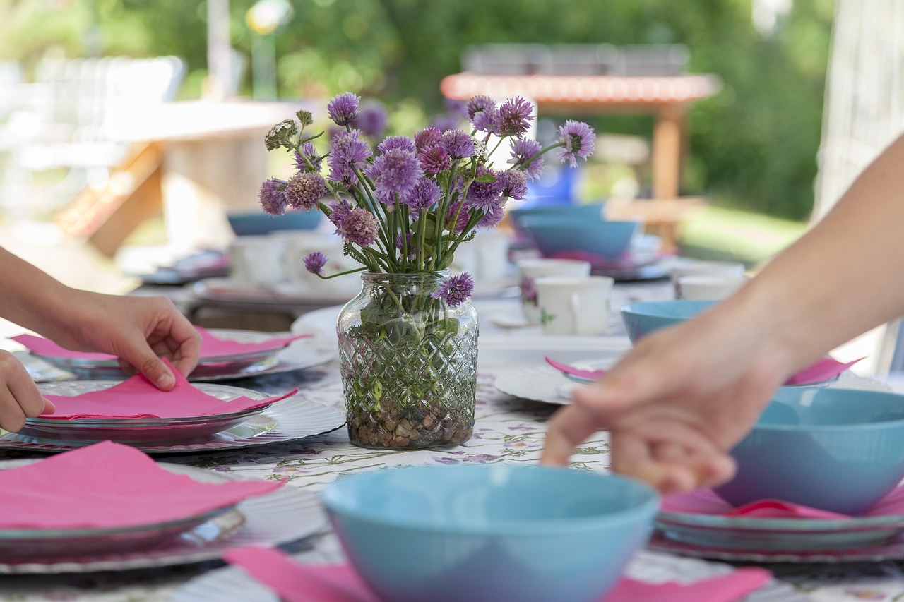 Hands setting an outdoor table with pink napkins, blue bowls, and a vase of purple chive flowers.