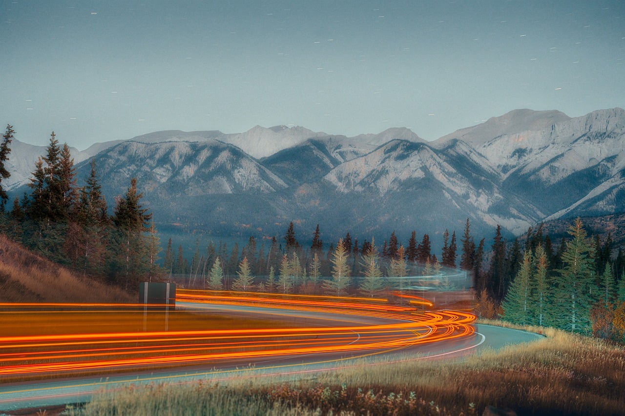 Winding road at night with orange light trails from vehicles, surrounded by pine trees and mountains.