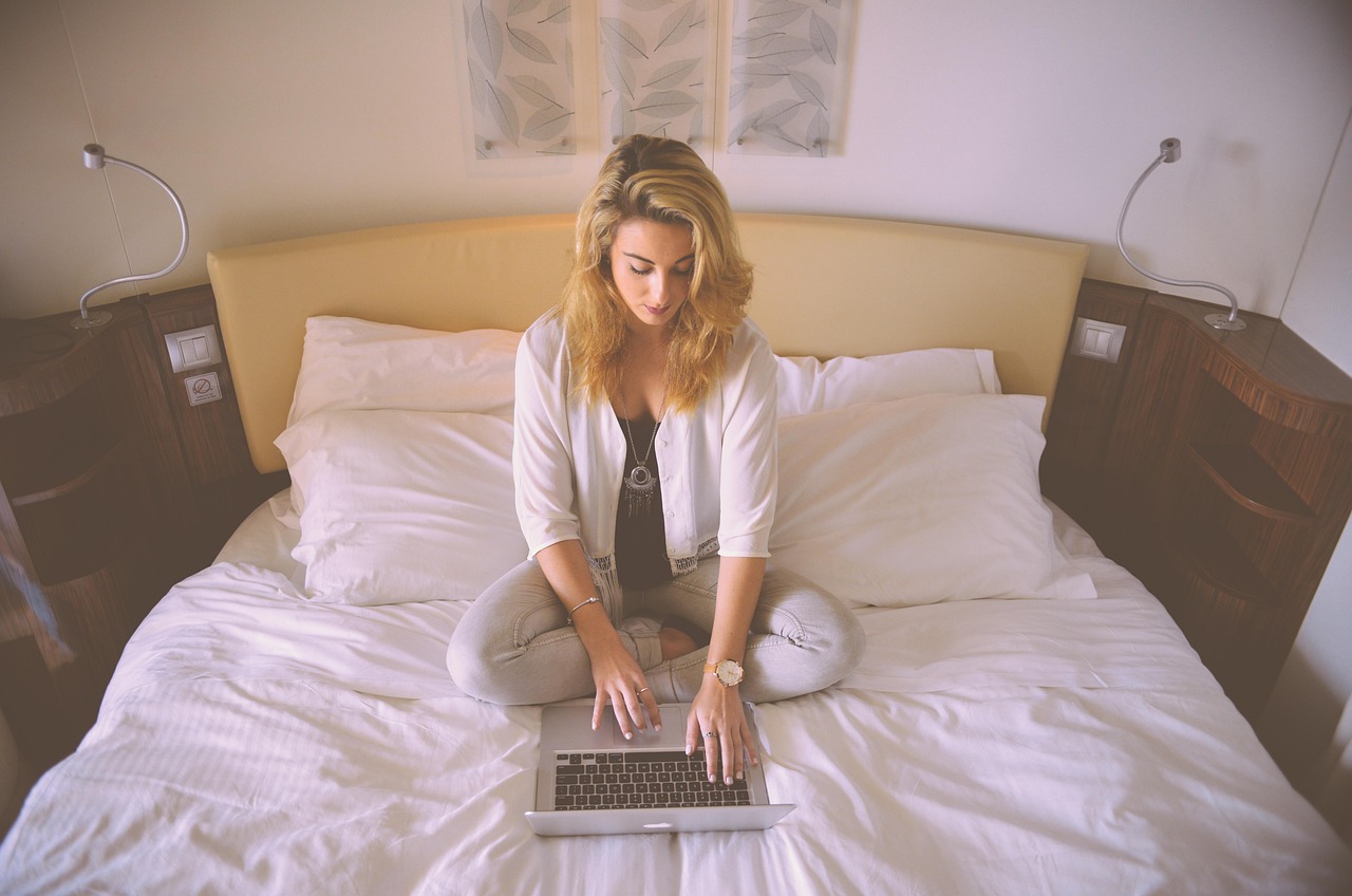 Young woman sitting cross-legged on a bed, typing on a silver laptop.