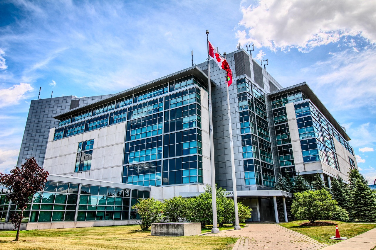 Modern government building with many windows and a Canadian flag flying on a flagpole under a blue sky.