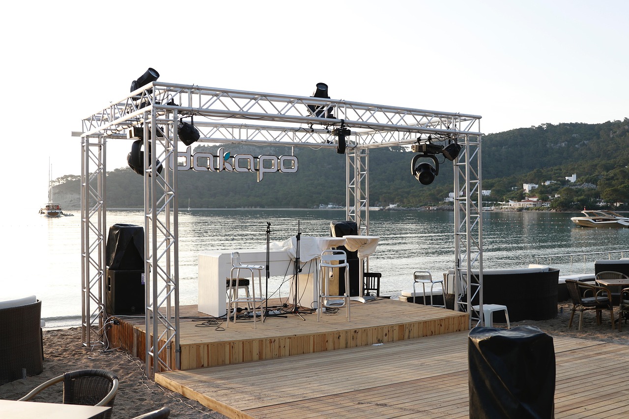 An outdoor stage with metal trussing, speakers, and lights on a sandy beach next to water, with hills and boats in the background.