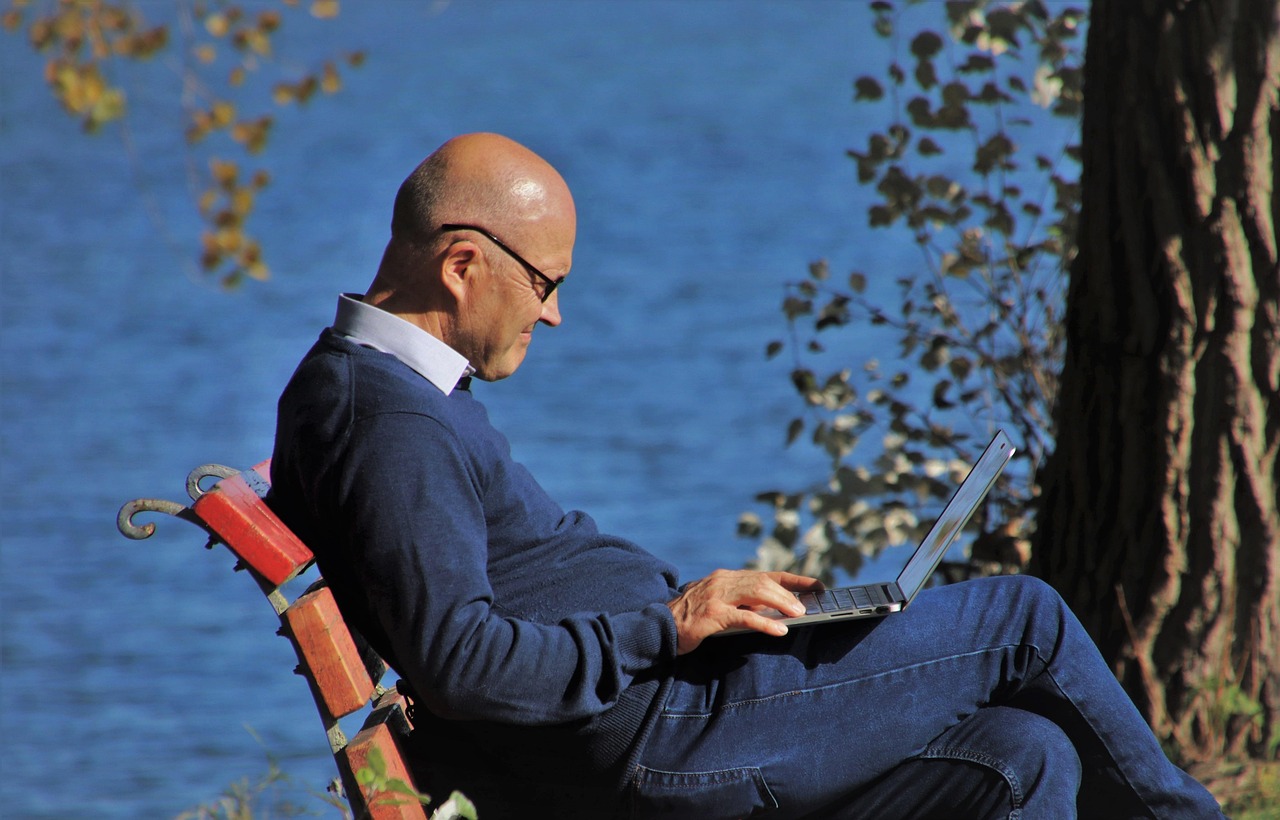 Working remotely from Canada on an eTA: a man with a laptop on a red bench by a lake in autumn sunlight